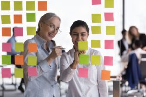 Younger and older woman working on collaborative project, writing ideas on sticky notes attached to transparent wall