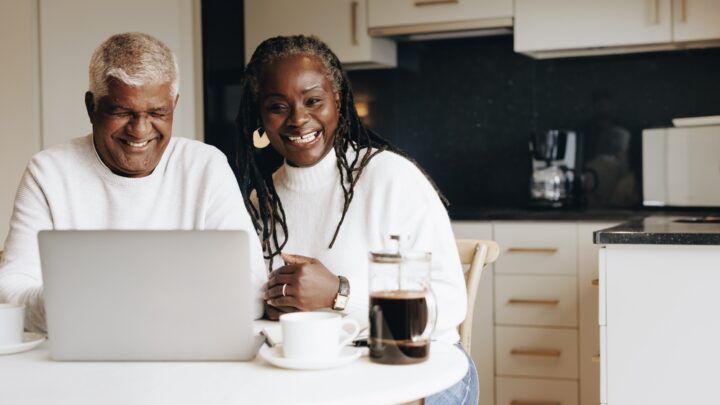 Older male and female couple sitting in their kitchen while using a laptop.